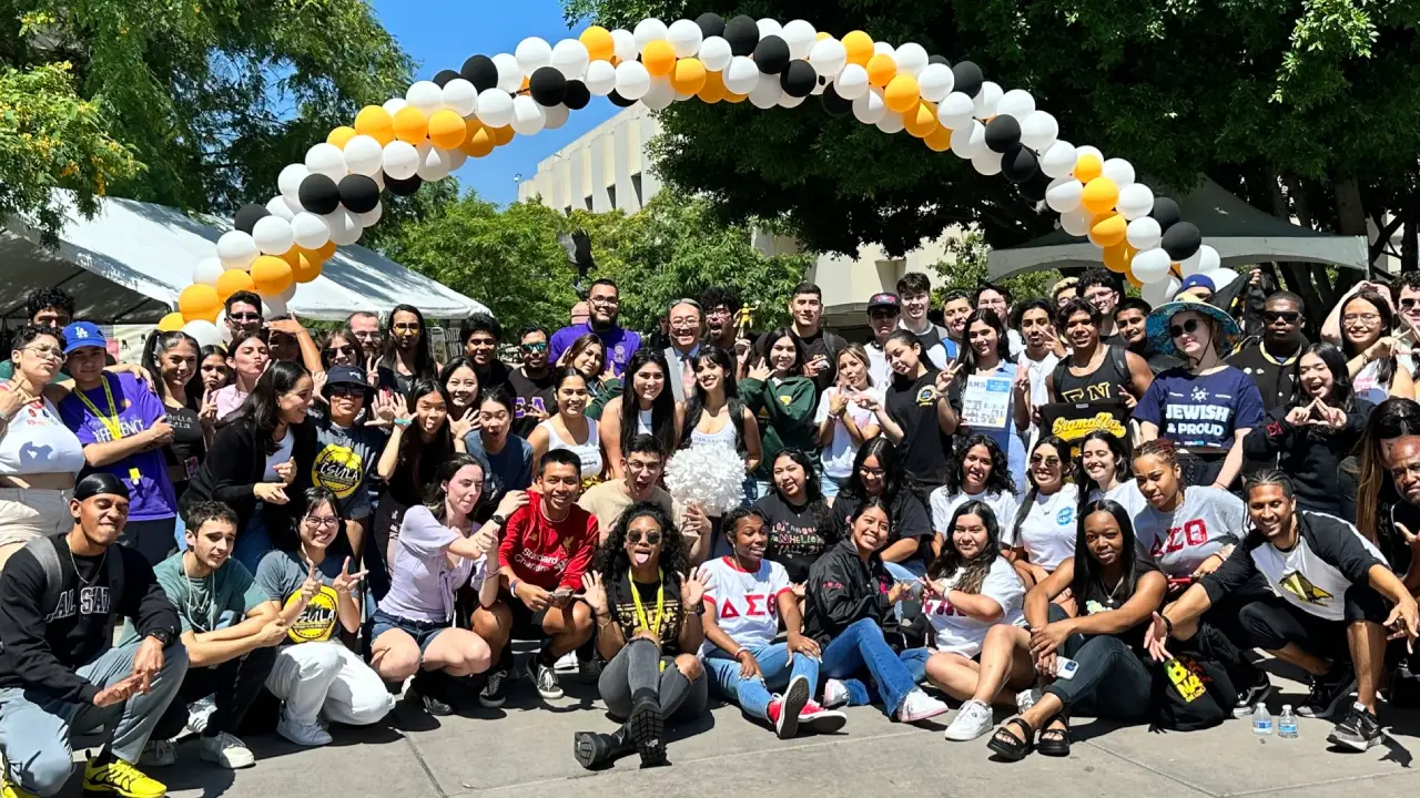 Large group of students gathered outdoors under an arch of black, white, and gold balloons
